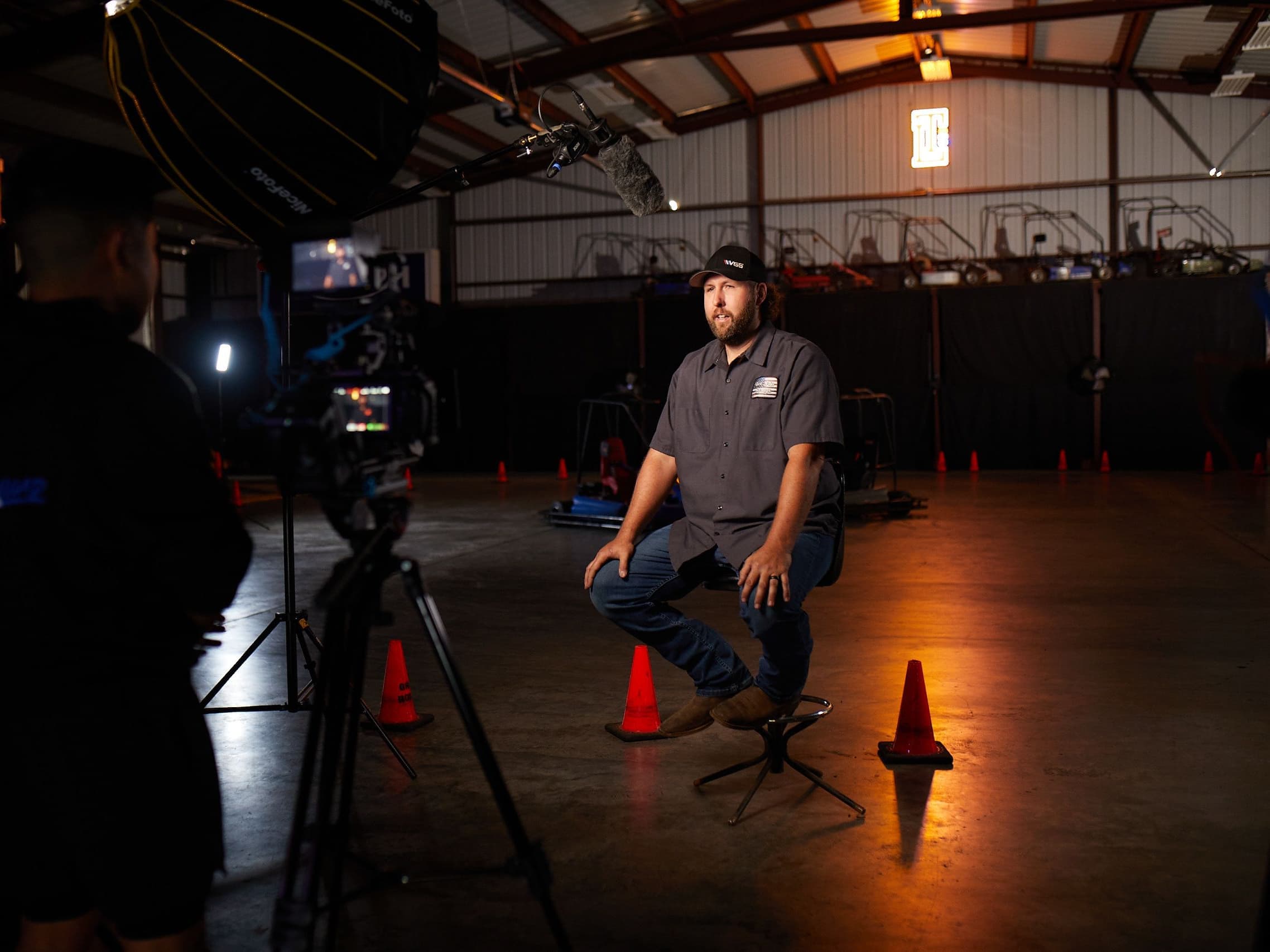 Interview being filmed in a lit hangar: subject on a stool, camera and softbox in the foreground